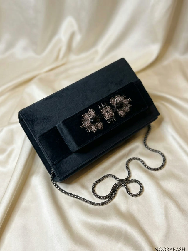 A black velvet clutch bag with three floral embroidered details on the front flap and a silver chain strap, displayed on a draped cream satin fabric background.
