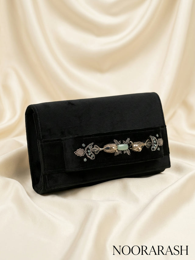 A black velvet clutch bag with an ornate silver-toned filigree and light green stone embellishment across the front flap, displayed on draped cream satin fabric. The brand name 'NOORARASH' is printed in the bottom right corner.