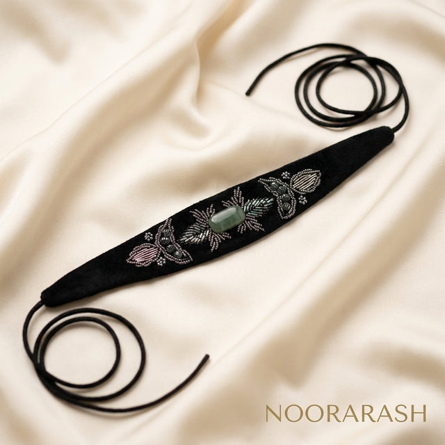 Black velvet Zinat choker necklace with silver, pink, and green floral embroidery and a central green stone, laid flat with long cord ties on cream satin fabric.