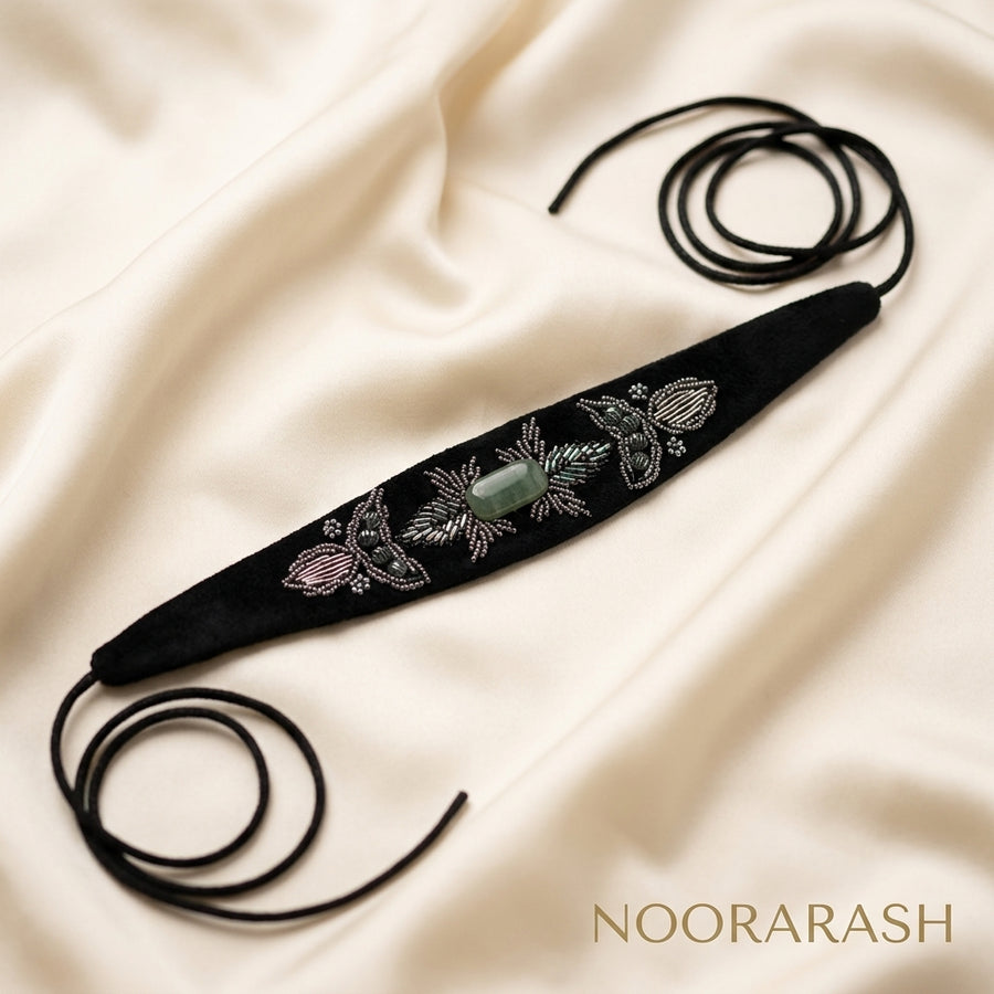 Black velvet Zinat choker necklace with silver, pink, and green floral embroidery and a central green stone, laid flat with long cord ties on cream satin fabric.