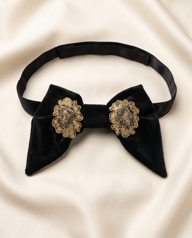 A black velvet bow tie with ornate gold floral embroidery on the front, displayed on a draped cream satin fabric background.