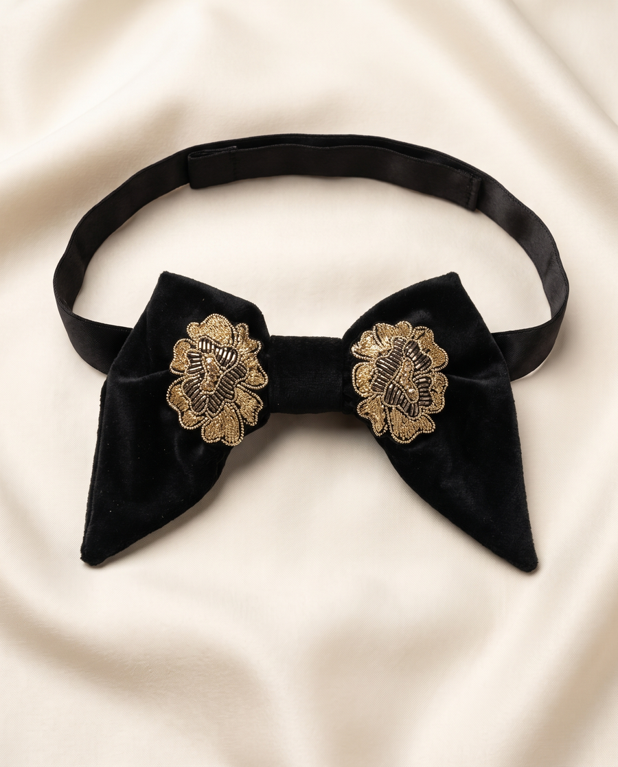 A black velvet bow tie with ornate gold floral embroidery on the front, displayed on a draped cream satin fabric background.