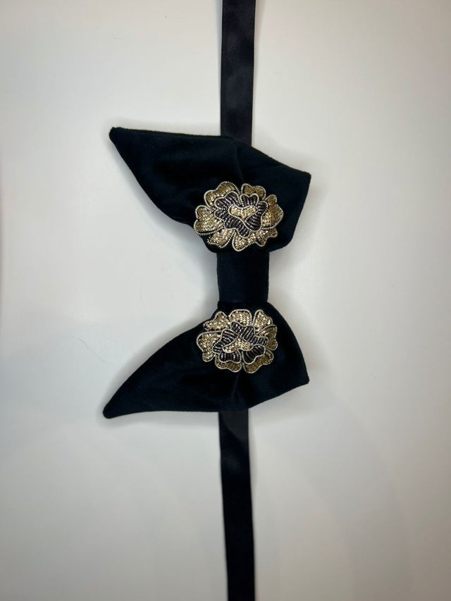 A black velvet bow tie with gold embroidered floral details, hanging against a plain white background.