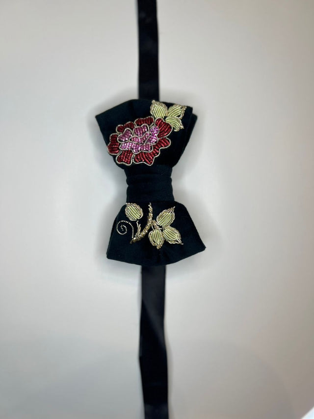 A black velvet bow tie with gold embroidered leaves and a red and pink floral motif on the upper loop, hanging against a plain light background.