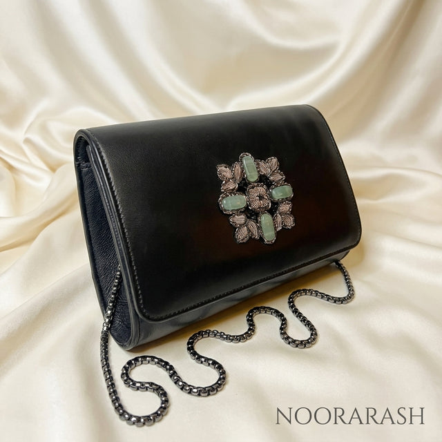 A black leather clutch bag with a decorative floral brooch featuring light green and pink stones, resting on a draped cream satin fabric. A black chain strap is draped in front of the bag.