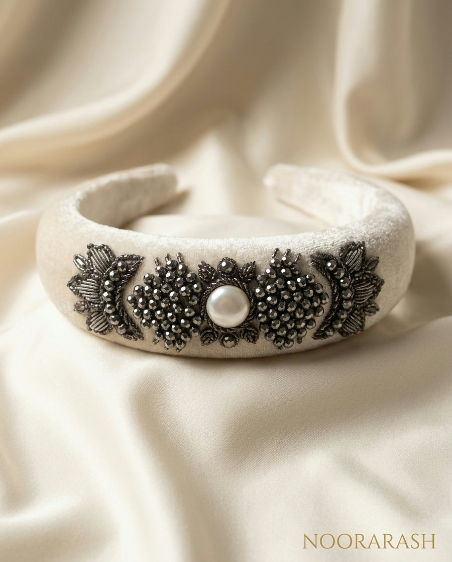 A wide cream-colored velvet headband with intricate black beaded floral and leaf patterns and a central white pearl, displayed on draped cream satin fabric.