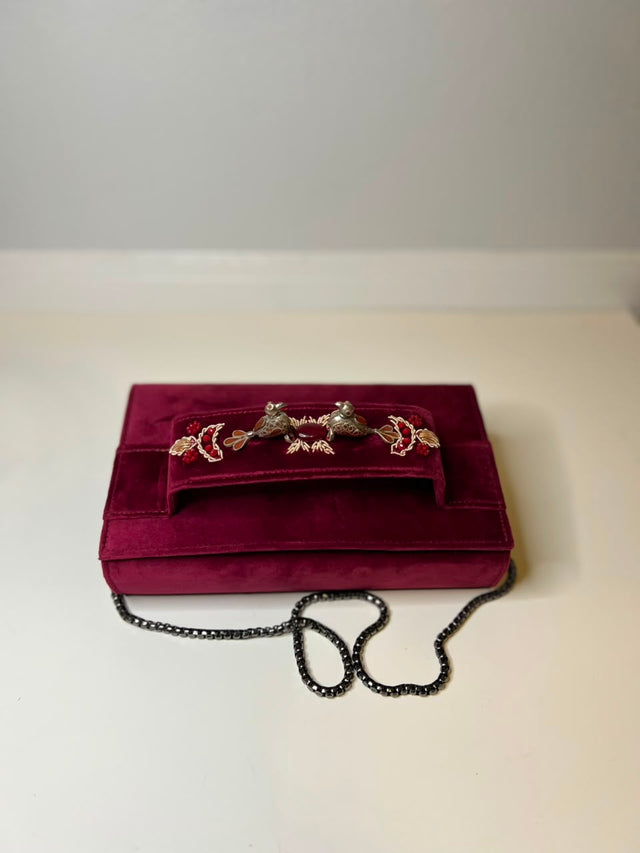 A deep magenta velvet clutch purse with a decorative silver-tone metal handle embellished with red and green crystals, resting on a light beige surface with a black chain strap.