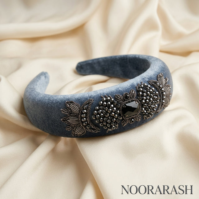 A wide blue velvet headband with ornate silver floral embroidery and a central black rhinestone, displayed on draped cream satin fabric. The brand name NOORARASH is printed in the bottom right corner.