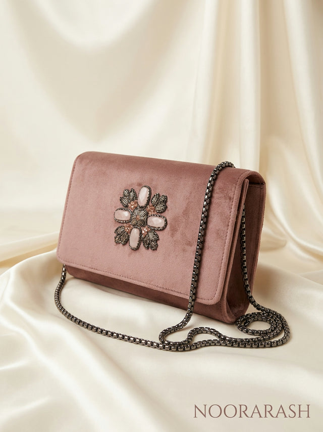 A dusty rose velvet clutch bag with a dark metal chain strap and a decorative floral embellishment on the front flap, displayed on draped cream satin fabric. The brand name 'NOORARASH' is visible in the bottom right corner.