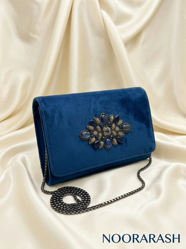 A royal blue velvet clutch bag with a large jeweled floral embellishment on the front flap and a black chain strap, displayed on draped cream satin fabric. The brand name 'NOORARASH' is visible in the bottom right corner.