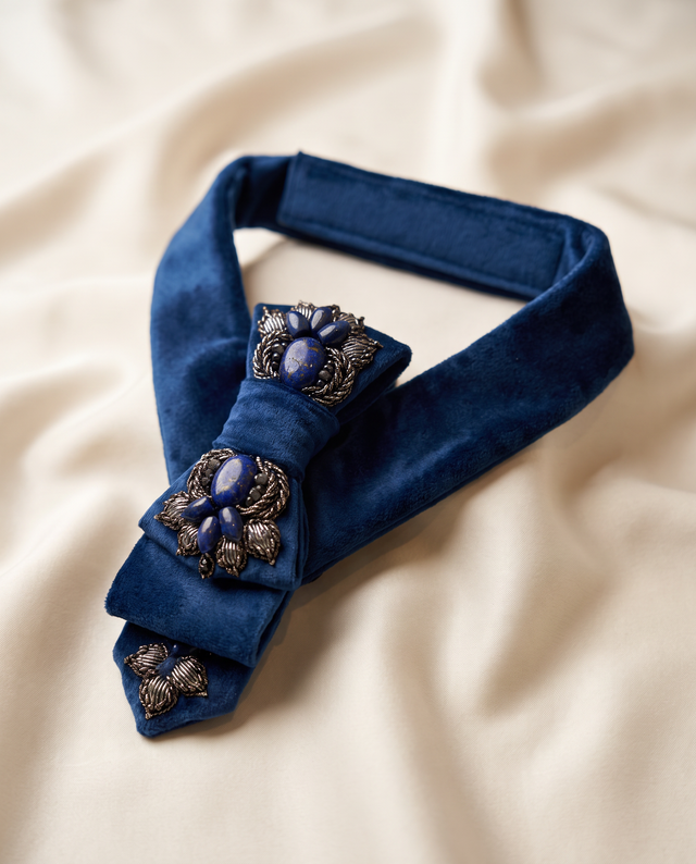 A dark blue velvet bow tie with ornate silver-tone floral brooch embellishments, laid flat on a cream-colored draped fabric background.