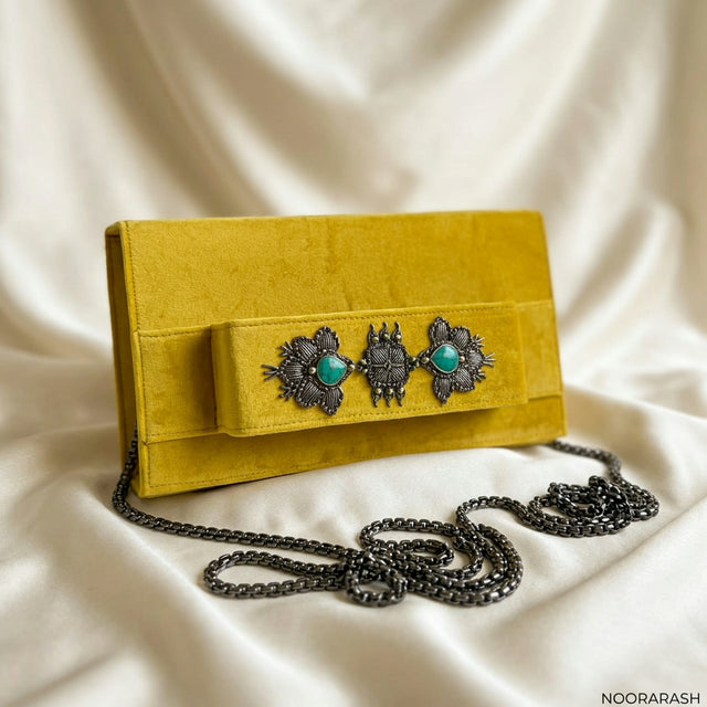 A mustard yellow suede clutch bag with a dark gunmetal chain strap and ornate metal embellishments on the front flap, resting on draped cream satin fabric.
