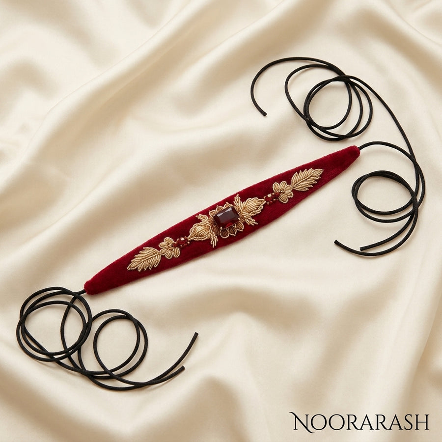 Red velvet Zinat choker necklace with gold floral embroidery and a central brown gemstone, laid flat with long cord ties on cream satin fabric.