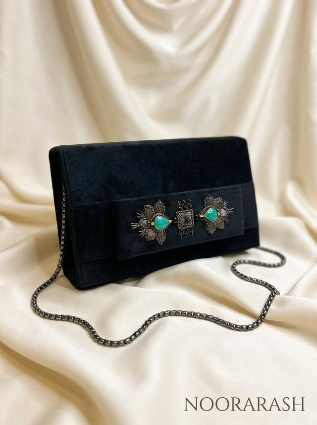 A black velvet clutch bag with a silver-tone chain strap and ornate turquoise and silver decorative hardware on the front flap, displayed on draped cream satin fabric. The brand name 'NOORARASH' is printed in the bottom right corner.