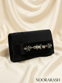 A black velvet clutch bag with an ornate silver-toned filigree and light green stone embellishment across the front flap, displayed on draped cream satin fabric. The brand name 'NOORARASH' is printed in the bottom right corner.