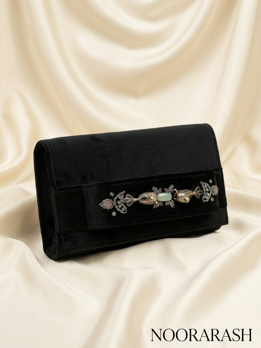 A black velvet clutch bag with an ornate silver-toned filigree and light green stone embellishment across the front flap, displayed on draped cream satin fabric. The brand name 'NOORARASH' is printed in the bottom right corner.