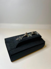A black velvet clutch purse with a silver-toned metal clasp featuring a floral design, resting on a light beige surface against a neutral gray background.