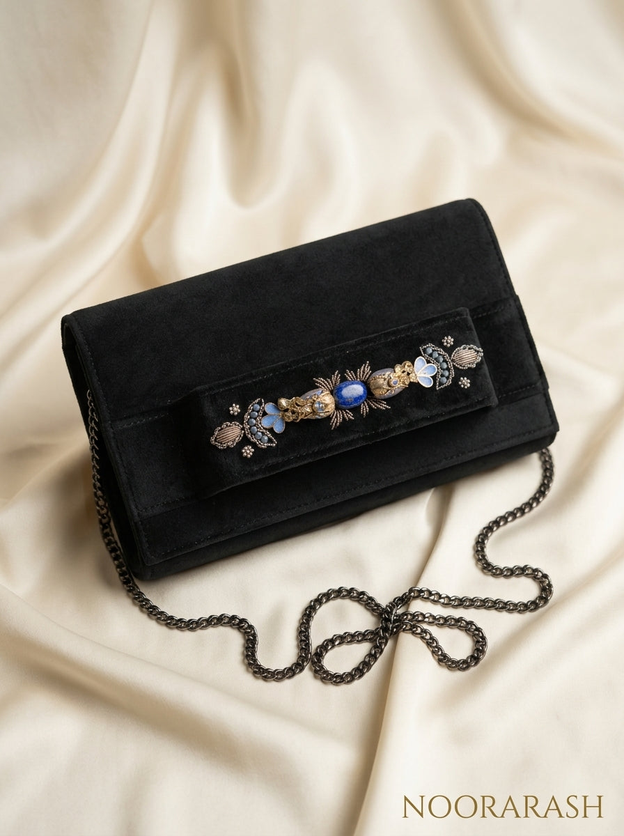 A black velvet clutch bag with an ornate decorative embellishment across the front flap, featuring blue and gold details, resting on draped cream-colored satin fabric. The brand name 'NOORARASH' is visible in the bottom right corner.