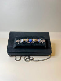 A black velvet evening bag with a decorative floral brooch featuring blue, white, and gold details, resting on a light beige surface with its silver chain draped in front.