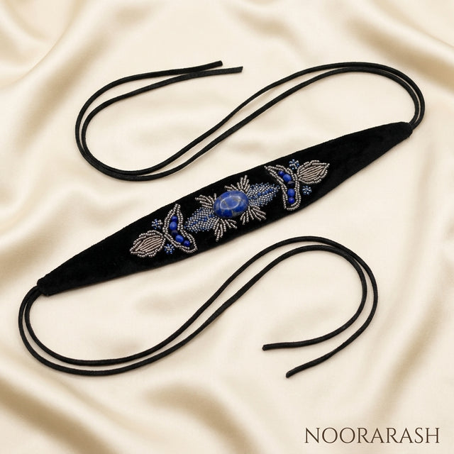 Black velvet Zinat choker necklace with blue, silver, and white floral beadwork, laid flat with long cord ties on draped cream satin fabric.