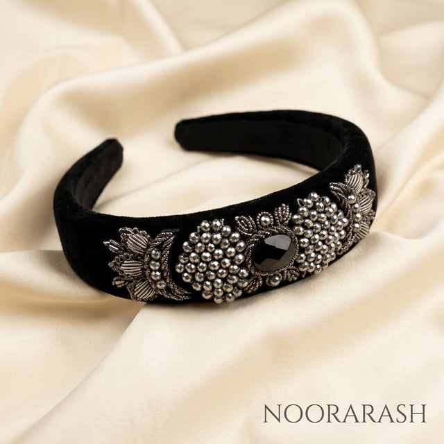 A black velvet wide headband with ornate silver floral embroidery and a central black faceted gemstone, displayed on draped cream satin fabric. The brand name NOORARASH is printed in the bottom right corner.