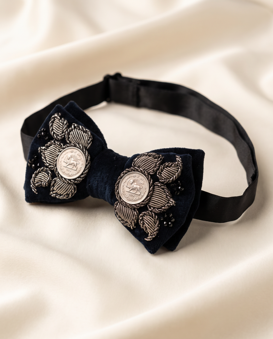 A dark navy blue velvet bow tie with silver embroidered floral details and circular silver medallions, displayed on a cream-colored satin fabric background.