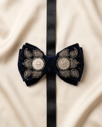 A dark navy blue velvet bow tie with intricate silver floral embroidery and two round silver medallion buttons, laid flat on a cream-colored fabric background with a vertical black ribbon running down the center.