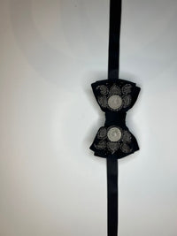 A black bow tie with silver filigree details and a silver coin in the center of each bow, hanging against a plain white background.