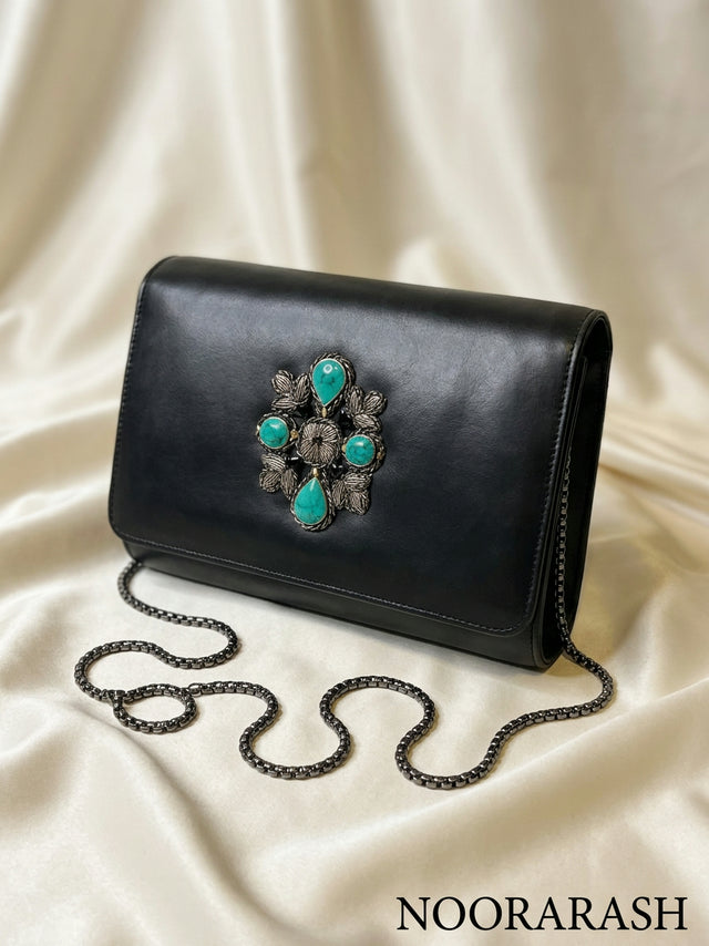 A black leather clutch bag with a large ornate turquoise and silver floral embellishment on the front, resting on draped cream satin fabric with a chain strap coiled in front. The brand name 'NOORARASH' is printed in the bottom right corner.