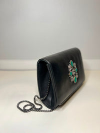 A black leather clutch bag with a silver chain strap and a decorative green and silver brooch on the front, photographed against a neutral light gray background.