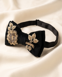 A black velvet bow tie with ornate gold embroidered floral and leaf motifs, displayed on draped cream-colored satin fabric.