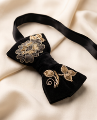 Black velvet bow tie with gold floral embroidery, displayed on a draped cream satin fabric background.