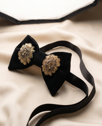 A black velvet bow tie with ornate gold floral brooch details on the front, displayed on a beige satin fabric background.