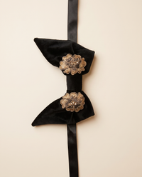 A black velvet bow tie with two ornate gold floral brooch accents on the center, attached to a long black ribbon, shown against a solid light beige background.