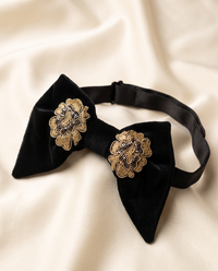 A black velvet bow tie with ornate gold embroidered floral details on the front, displayed on a draped cream satin fabric background.