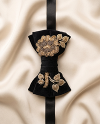 A black velvet bow tie with intricate gold and silver floral embroidery, displayed on a draped beige satin fabric background.