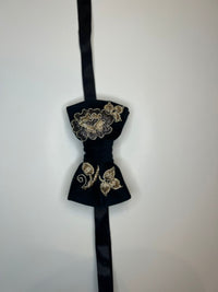 A black velvet bow tie with gold floral embroidery, hanging against a plain off-white wall.