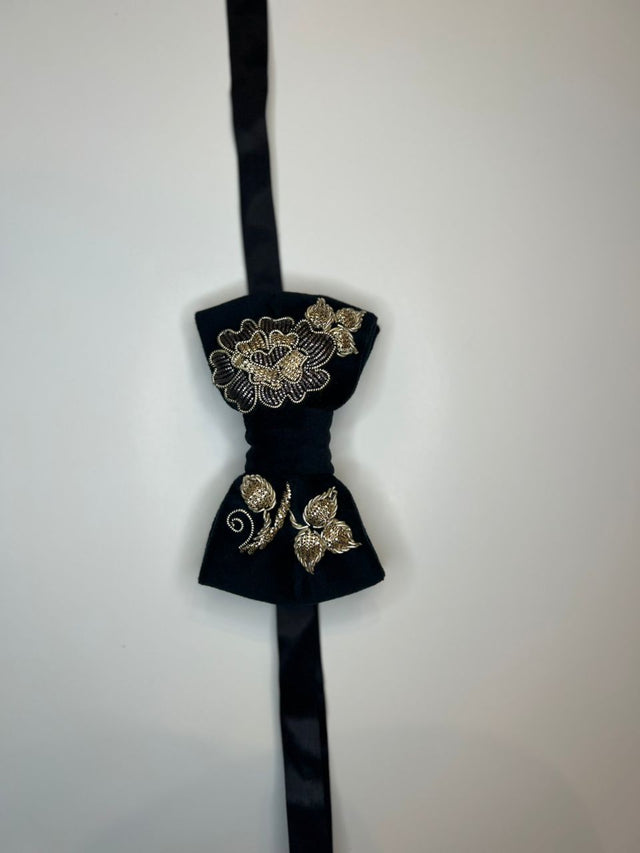 A black velvet bow tie with gold floral embroidery, hanging against a plain off-white wall.