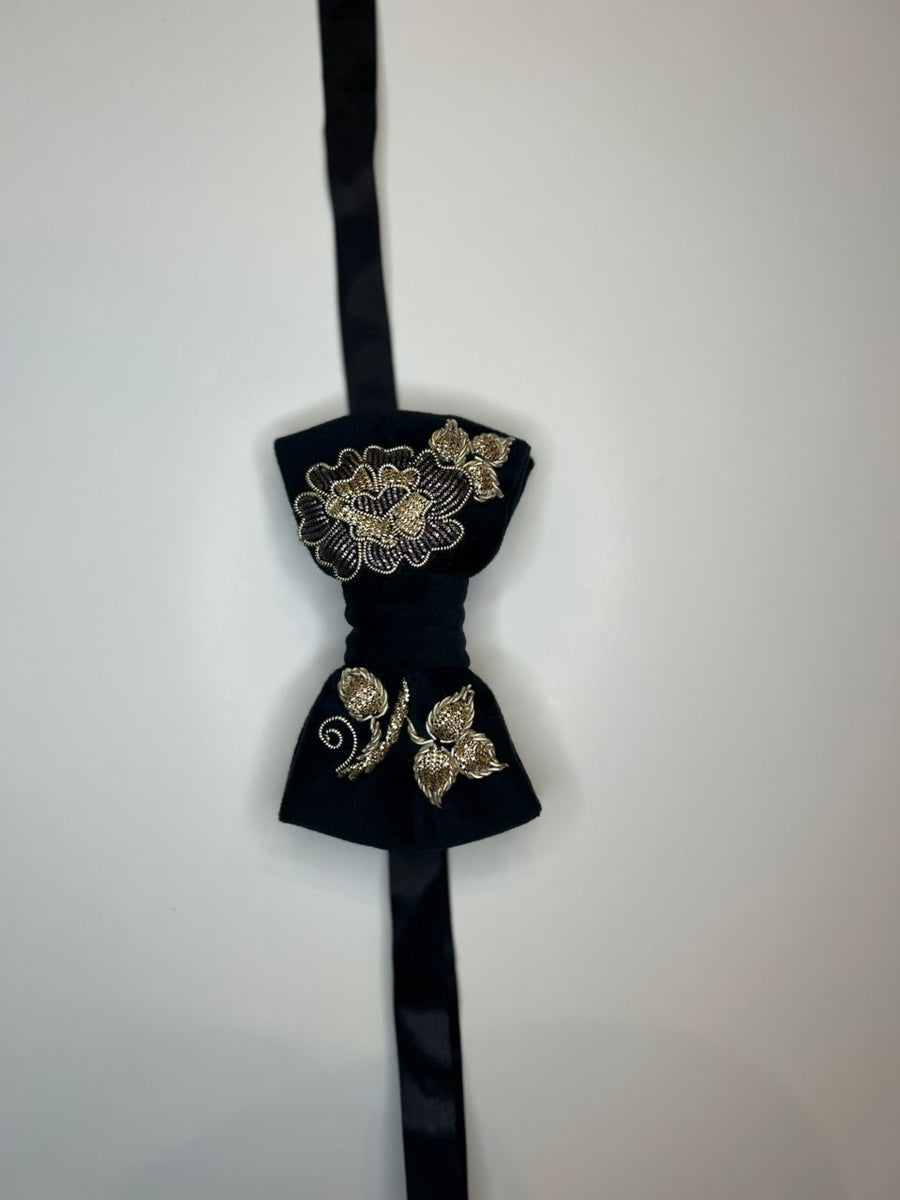 A black velvet bow tie with gold floral embroidery, hanging against a plain off-white wall.