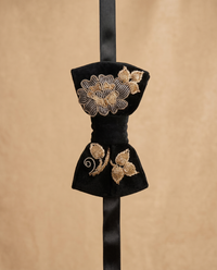 A black velvet bow tie with intricate gold floral embroidery, hanging on a black ribbon against a textured beige background.