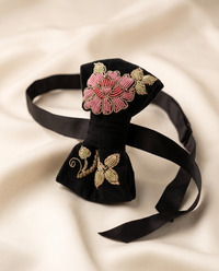 A black velvet bow tie with pink and gold floral Sermeh embroidery, displayed on a draped cream satin fabric background.
