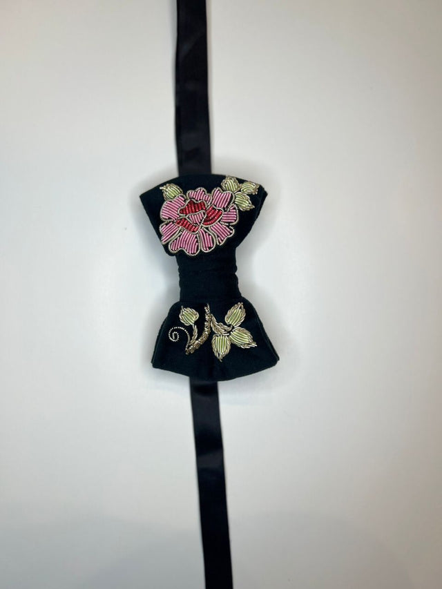 A black bow tie with intricate floral embroidery in pink, red, and gold on the front, hanging against a plain white background.