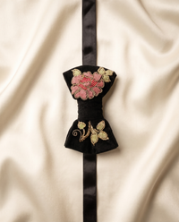 A black velvet bow tie with intricate floral embroidery in pink, red, and gold thread, tied with a black satin ribbon on a draped cream satin fabric background.