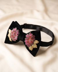 A black velvet bow tie with intricate floral embroidery in pink, red, and gold on both sides, displayed on a soft beige fabric background.