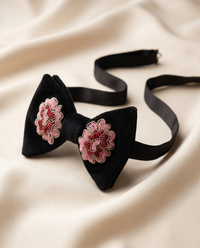 A black velvet bow tie with pink and white embroidered floral details on each wing, displayed on a draped cream satin fabric background.