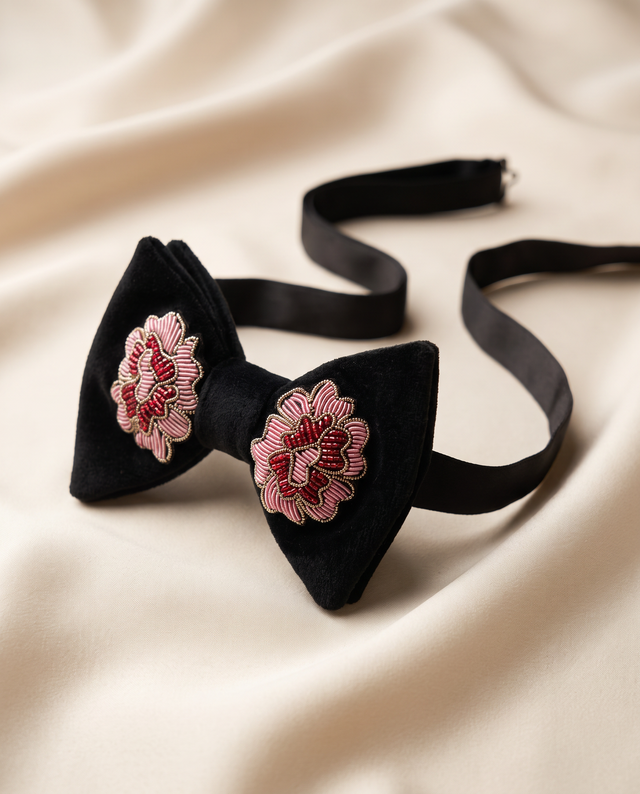 A black velvet bow tie with pink and white embroidered floral details on each wing, displayed on a draped cream satin fabric background.
