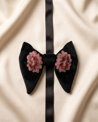 A black velvet bow tie with pink and red floral embroidery on each side, displayed on a cream-colored satin fabric background.