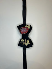 A black velvet bow tie with intricate floral embroidery, featuring a red and purple flower on the top bow and gold leaves on the bottom bow, attached to a black ribbon.