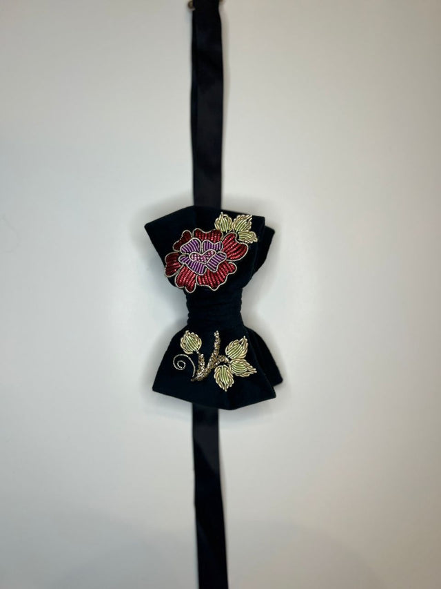 A black velvet bow tie with intricate floral embroidery, featuring a red and purple flower on the top bow and gold leaves on the bottom bow, attached to a black ribbon.
