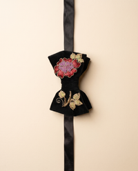 A black velvet bow tie with intricate floral embroidery in red, pink, and gold on both ends, suspended against a solid beige background.